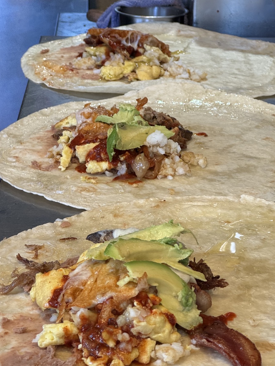 Breakfast burritos being assembled in a row with eggs, carnitas and avocado