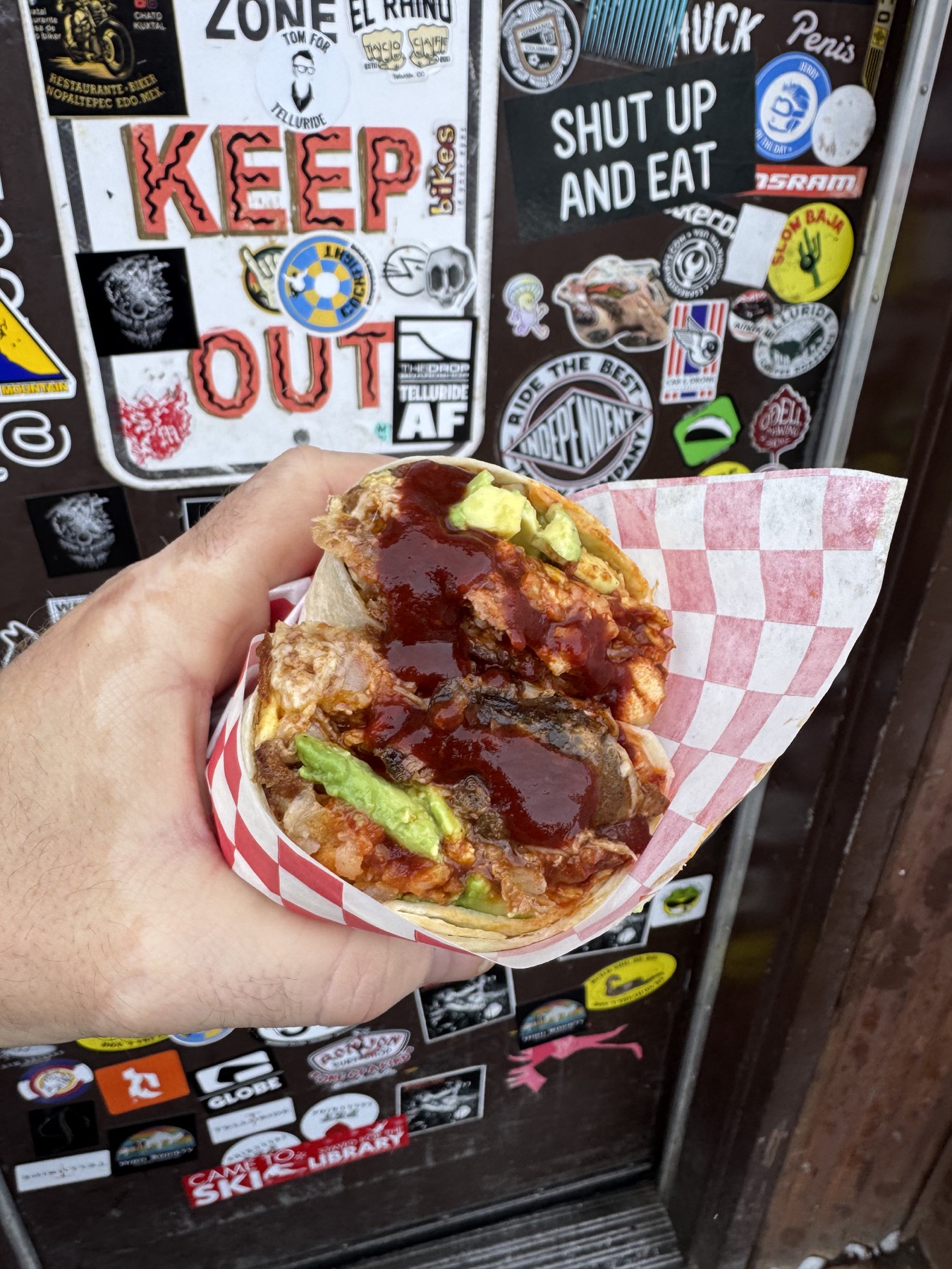 A saucy carnitas burrito held in front of El Rhino’s sticker-covered entrance