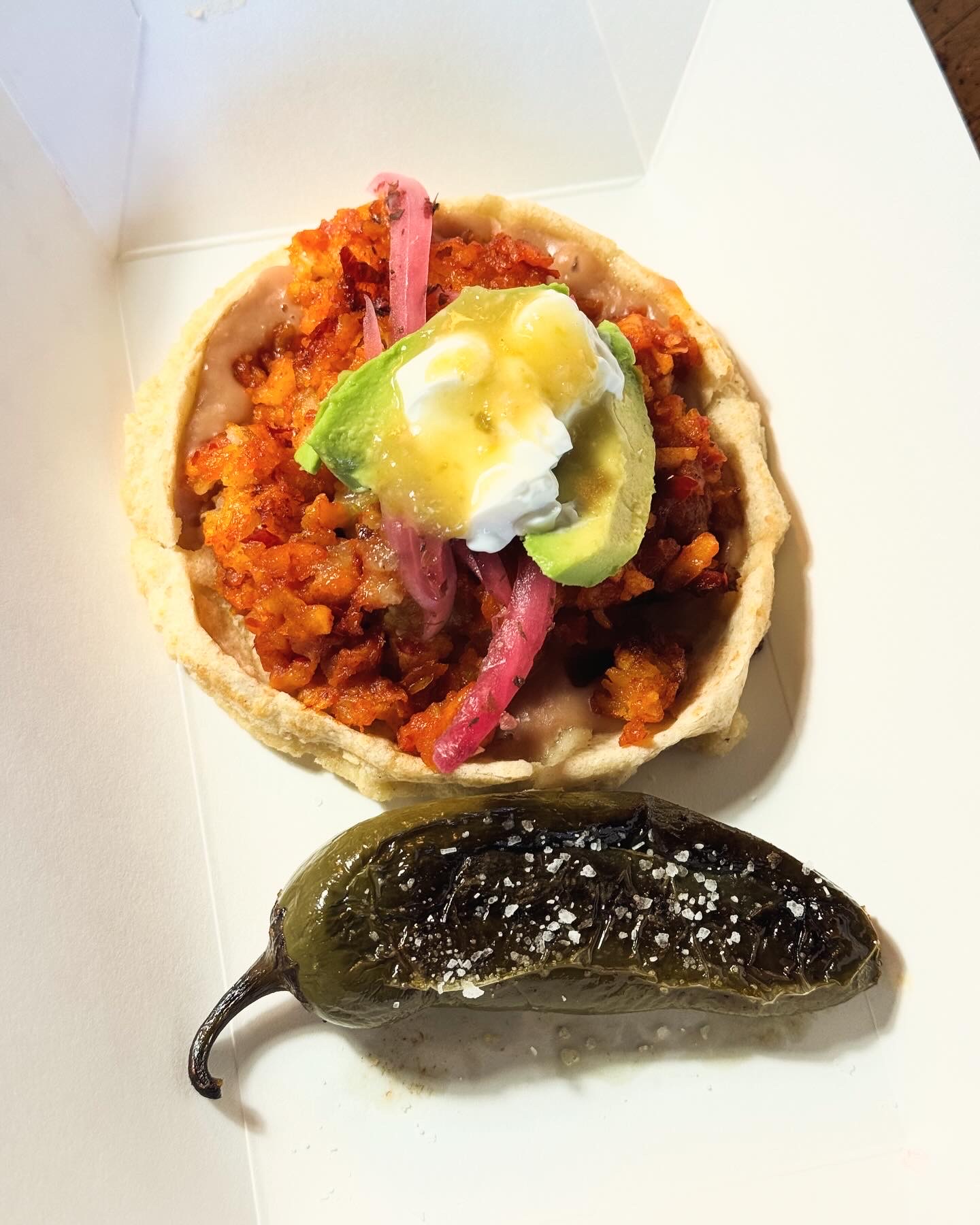 A colorful taco topped with avocado, crema, salsa and pickled onions beside a grilled jalapeño